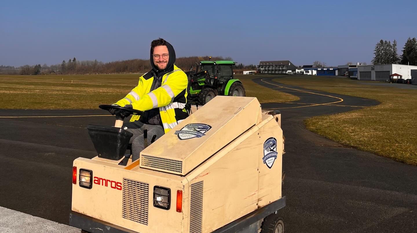 Vereinsmitglied bedient eine kompakte Kehrmaschine auf dem Rollweg, im Hintergrund Traktor und Flugplatzgebäude